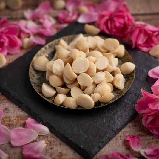 Macadamia Style 4 aus Kenia. Halbe und Viertel Kerne. Ansicht in Schale auf Schieferplatte mit Dekoration Rosa Blüten auf Holztisch