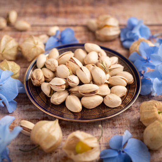Geröstete Pistazien ohne Salz aus den USA von nussmart. Ansicht in Schale auf Holztisch dekoriert mit blauen Blumenblüten