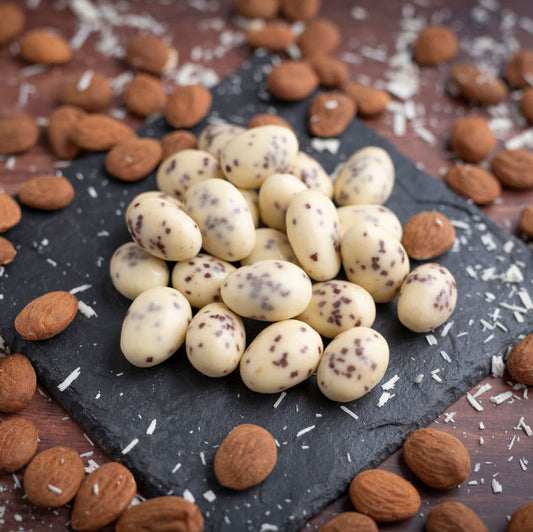nussmart Schoko Mandeln Straciatella. Schokolierte Mandeln mit weisser Schokolade und Schokoraspel. Dekoriert mit weissen Schokoladenraspeln und Mandeln auf Schieferplatte und dunkelbraunem Holztisch