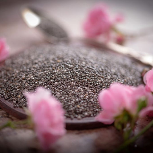 Chia Samen in einer Holzschale mit Löffel – umgeben von rosa Blüten auf rustikalem Holzhintergrund. Hochwertige Chiasamen als Superfood